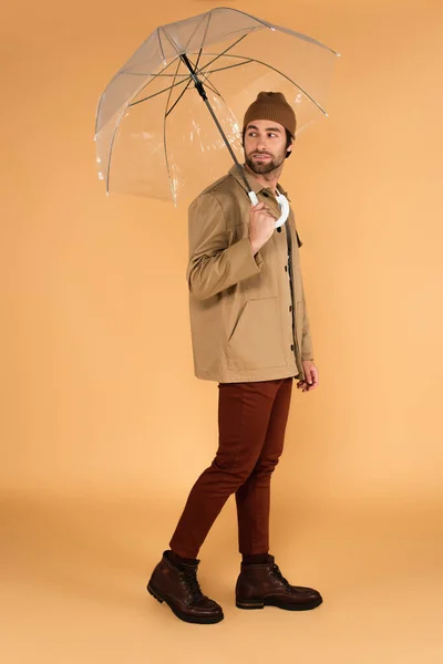 Молодий чоловік в куртці і коричневих штанах озирається назад під час ходьби під парасолькою на бежевому — стокове фото