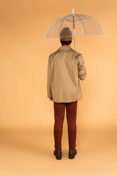 Back view of man in brown pants and jacket standing under transparent umbrella on beige — Stock Photo