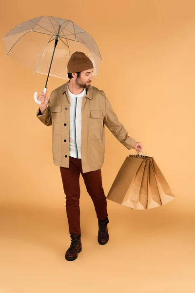 Mann in brauner Hose und Jacke trägt Einkaufstaschen unter Regenschirm auf beige — Stockfoto