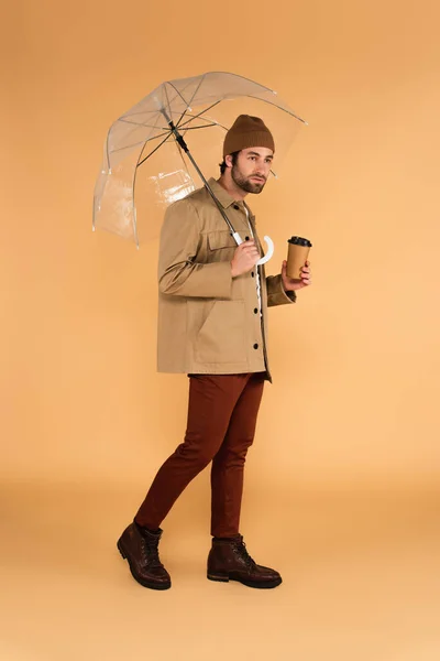 Full length view of trendy man walking with coffee to go under umbrella on beige — Stock Photo