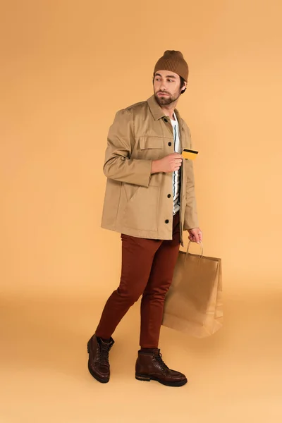 Чоловік в модному осінньому вбранні, що йде з кредитною карткою і сумками на бежевому — стокове фото