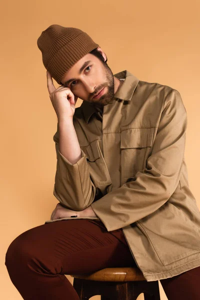 Man in trendy autumn clothes looking at camera while sitting with hand near head isolated on beige - foto de stock