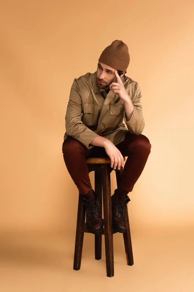 Молодий чоловік у стильних чоботях і куртці, що сидить на дерев'яному табуреті на бежевому — стокове фото