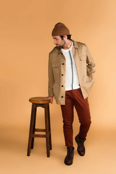 Stylish man in beanie, jacket and brown pants posing near wooden stool on beige — Foto stock