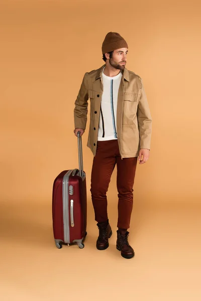Повнометражний вигляд стильного чоловіка з валізою, що дивиться в сторону на бежевому — стокове фото