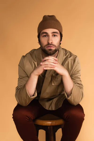 Bearded man in stylish jacket and beanie sitting with clenched hands isolated on beige — Foto stock