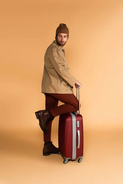 Мужчина в коричневых брюках, сапогах и куртке, стоящий рядом с чемоданом на бежевом — стоковое фото