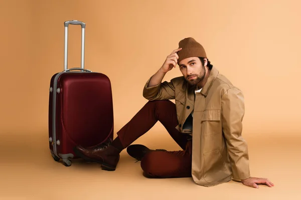 Young bearded man in trendy autumn clothes sitting on floor near suitcase on beige — Fotografia de Stock