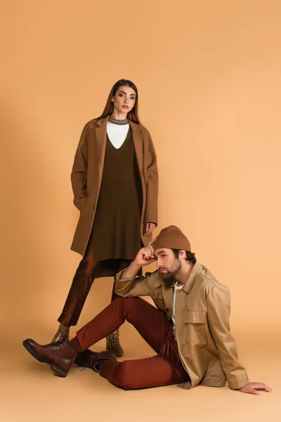 Young and stylish woman looking at camera near trendy man sitting on beige background — Photo de stock
