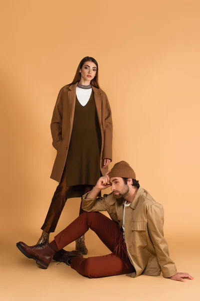 Young and stylish woman looking at camera near trendy man sitting on beige background — Stock Photo