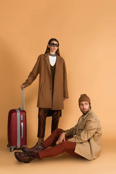 Man in jacket and beanie sitting near stylish woman and suitcase on beige background — Foto stock