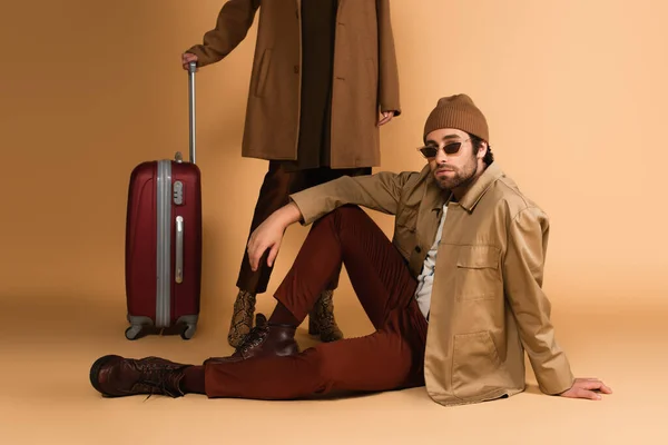 Trendy man in sunglasses sitting near woman and suitcase on beige background — Fotografia de Stock