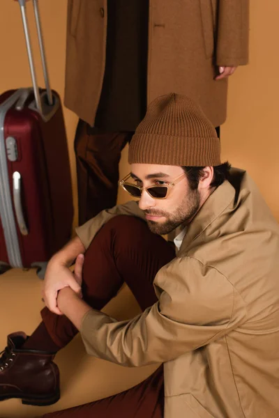 Мужчина в модных солнцезащитных очках, сидящий рядом с размытой женщиной с чемоданом бежевого цвета — стоковое фото