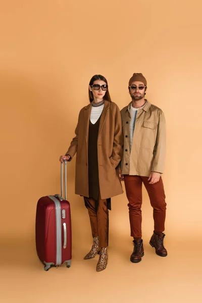 Young fashionable couple in sunglasses near suitcase on beige background — Fotografia de Stock