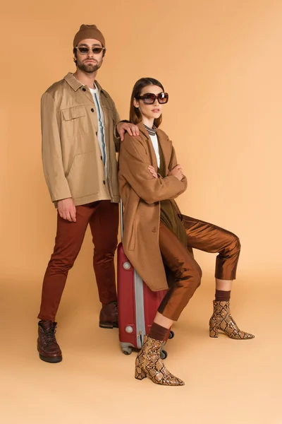 Stylische Frau auf Koffer neben trendigem Mann auf beigem Hintergrund — Stockfoto