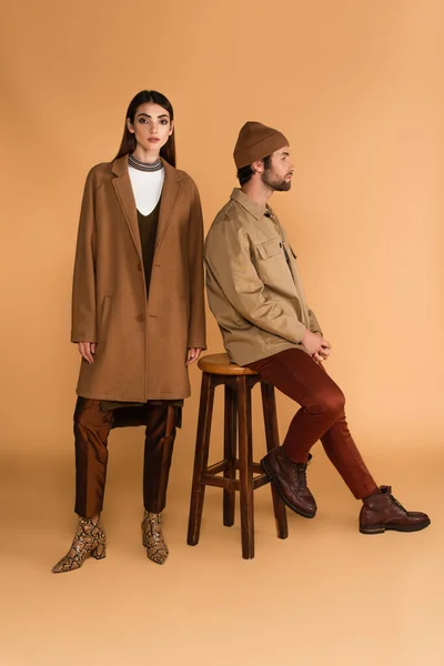 Young woman in trendy coat near man sitting on stool in jacket and beanie on beige — Fotografia de Stock