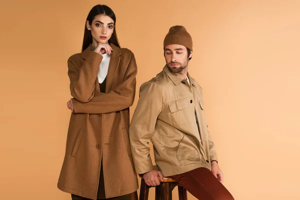 Woman in brown coat standing near stylish man sitting on stool isolated on beige — Foto stock