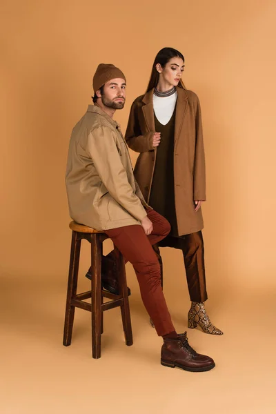 Trendy man in jacket and beanie sitting on high stool near stylish woman on beige background — Fotografia de Stock