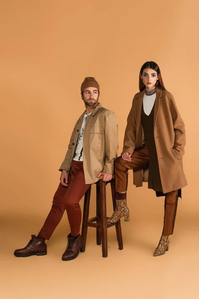 Woman with hand in pocket of stylish coat near trendy man sitting on high stool on beige background — Fotografia de Stock