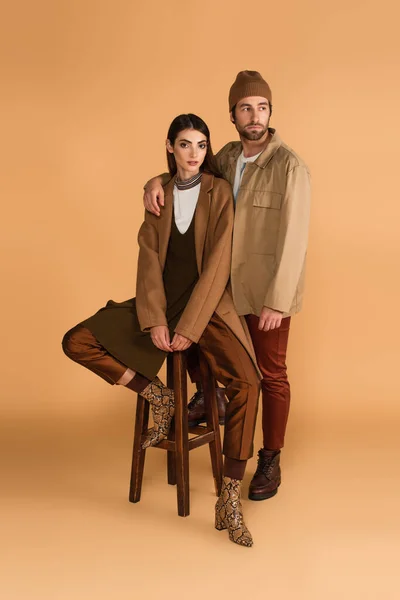 Stylish man hugging shoulder of fashionable woman sitting on high stool on beige background — Stock Photo