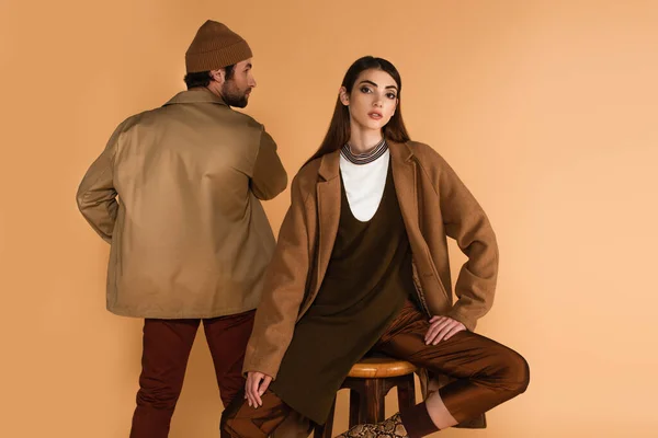 Rückansicht eines Mannes in Jacke neben einer stilvollen Frau, die isoliert auf einem Hocker in Beige sitzt — Stockfoto