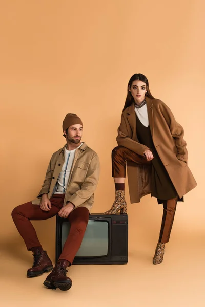Женщина в стильном осеннем пальто смотрит на камеру рядом с модным мужчиной, сидящим на винтажном телевизоре на бежевом фоне — стоковое фото