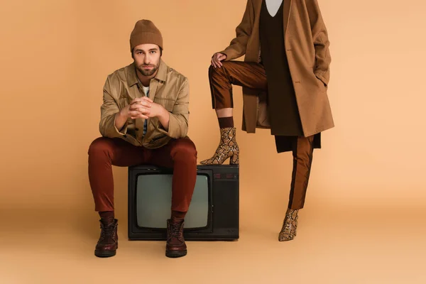 Молодой человек в осенней куртке и шапочка сидит на винтажном телевизоре рядом с женщиной в стильных сапогах на бежевом фоне — стоковое фото