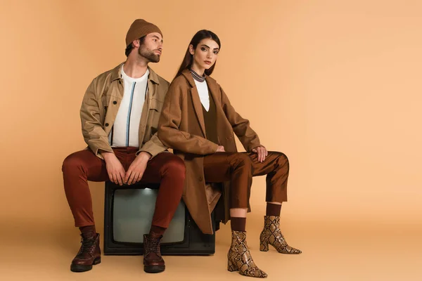 Young couple in fashionable autumn outfit sitting on vintage tv set on beige background — Foto stock