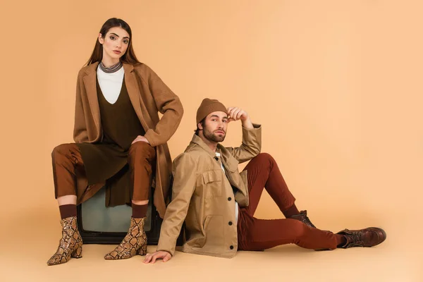 Trendy woman sitting on vintage tv set near stylish man on floor on beige background — Stock Photo