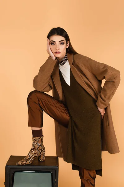 Fashionable woman stepping on retro tv set while posing with hand on hip isolated on beige — Stock Photo