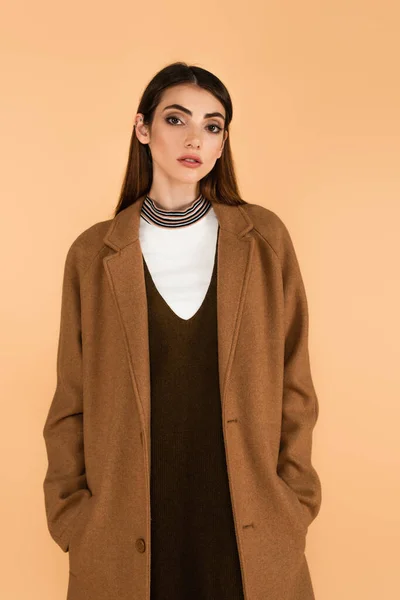 Pretty young woman holding hands in pockets of brown coat isolated on beige — Stock Photo