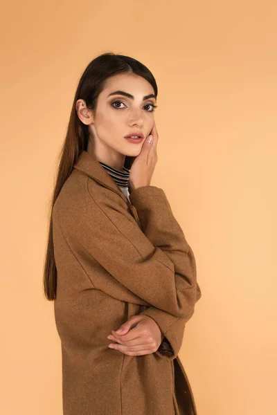Young woman in brown coat holding hand near face while looking at camera isolated on beige - foto de stock