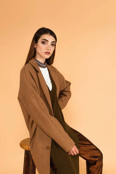 Trendy woman in brown coat sitting on stool with hand on hip isolated on beige — Foto stock