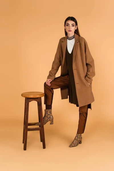 Trendy woman holding hand in pocket of coat while posing near high stool on beige — Stock Photo