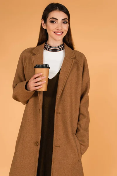 Happy woman with coffee to go standing with hand in pocket of stylish coat isolated on beige — Foto stock
