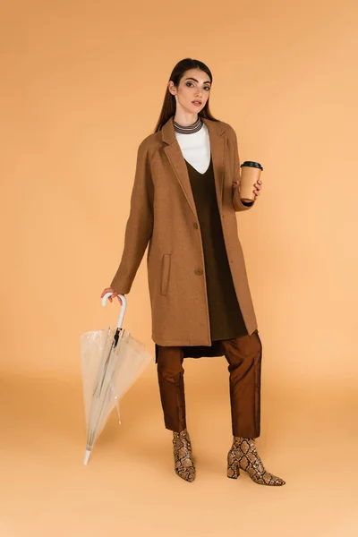 Брюнетка в модной осенней одежде позирует с бумажной чашкой и зонтиком на бежевом — стоковое фото
