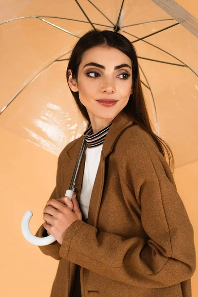 Позитивная молодая женщина в коричневом пальто глядя в сторону под зонтиком, изолированным от бежевого — стоковое фото
