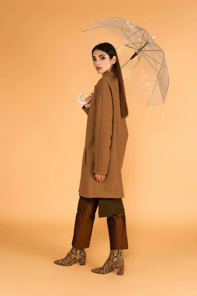 Pretty woman in brown coat and leather boots standing under transparent umbrella on beige background — Photo de stock