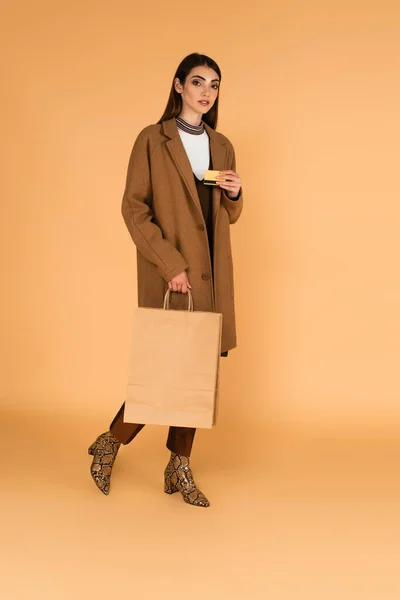 Brunette woman in stylish autumn outfit walking with credit card and shopping bag on beige — Stock Photo