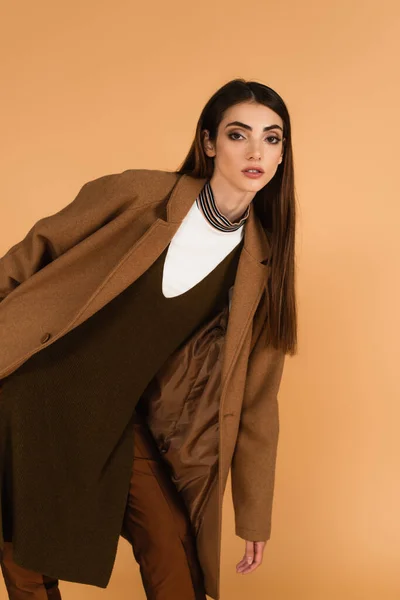 Young brunette woman in brown coat looking at camera while posing isolated on beige — Fotografia de Stock