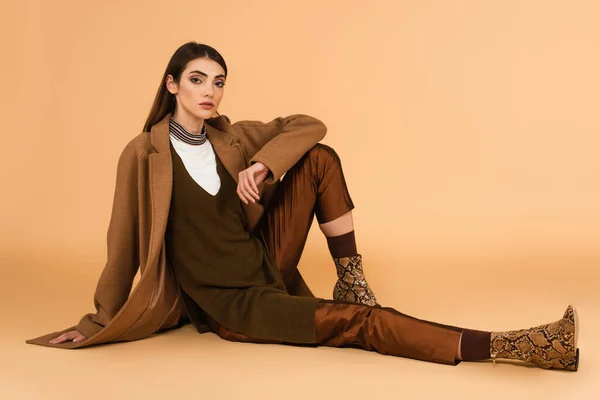 Young woman in brown coat, satin pants and leather shoes sitting on beige background — Foto stock