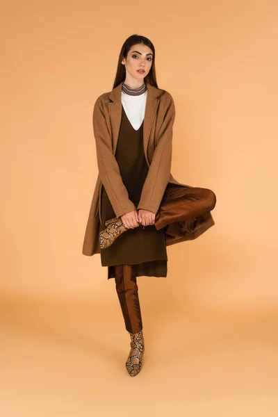 Fashionable woman in brown coat and leather boots posing on one leg on beige — Stock Photo