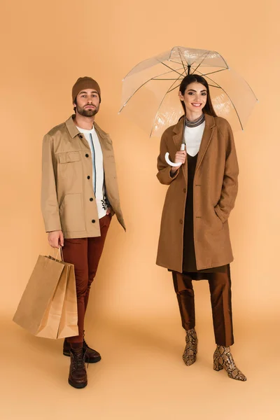 Smiling woman under transparent umbrella near trendy man with shopping bags on beige background — Fotografia de Stock