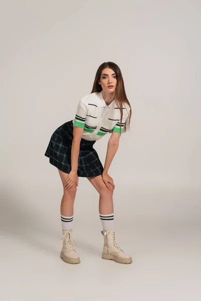Trendfrau in Polo-T-Shirt, kariertem Rock und Lederstiefeln blickt in die Kamera auf grauem Hintergrund — Stockfoto