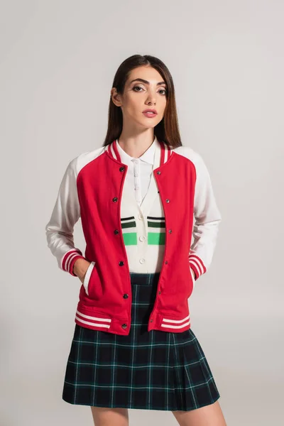Brunette woman in plaid skirt posing with hands in pockets of jacket isolated on grey - foto de stock