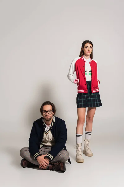 Stylish man sitting with crossed legs near stylish woman posing with hands in pockets on grey background — Stock Photo