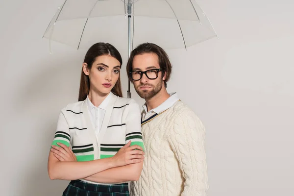 Fashionable couple looking at camera under transparent umbrella on grey background — Foto stock