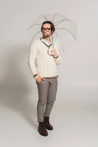 Full length view of stylish man standing under umbrella with hand in pocket on grey — Fotografia de Stock