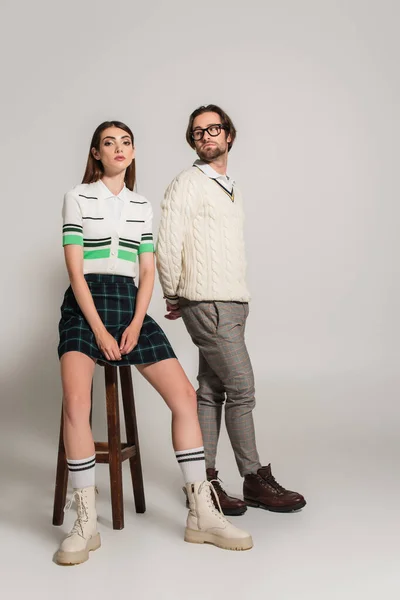 Woman in plaid skirt and boots sitting on high stool near trendy man on grey background — Fotografia de Stock