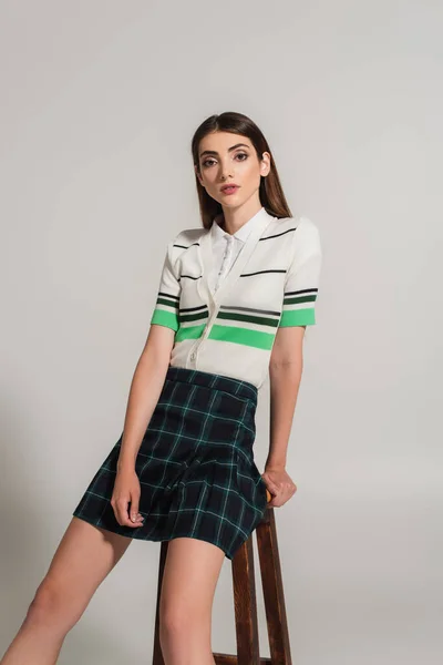 Pretty woman in striped polo t-shirt and plaid skirt looking at camera on grey background - foto de stock